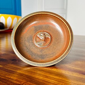 May include: A small, round, brown ceramic dish with a raised, embossed design of a bird in the center. The dish has a dark brown rim and a slightly textured surface.