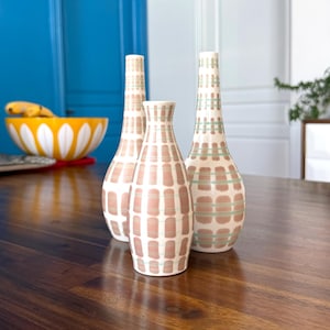 May include: Three ceramic vases with a mid-century modern design. The vases are white with a grid pattern in shades of pink and green. They are arranged on a dark wood table. The background includes a blue wall and a white door.
