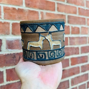 May include: A brown ceramic mug with a geometric design featuring stylised animals in a light brown colour. The mug has a black rim and a black geometric pattern around the top and bottom.
