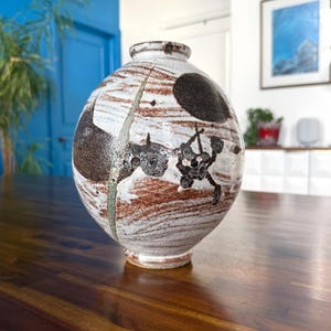 May include: A round ceramic vase with a wide body and narrow neck. The vase features a white base with brown and black abstract designs. A green line runs vertically down the side. The vase sits on a wooden surface.