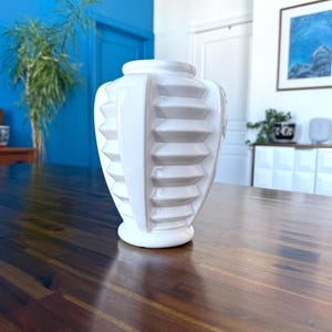 May include: A white ceramic vase with a geometric design. The vase has a wide base and a narrow neck. The body of the vase features a series of horizontal, stepped ridges. The vase is sitting on a dark wood table. The background includes a blue wall and a framed picture.