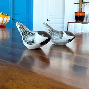 May include: Two ceramic bird figurines with black and white painted details. The birds are facing opposite directions and are sitting on a wooden surface.