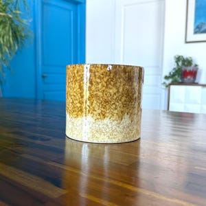 May include: A cylindrical ceramic vase with a speckled brown and tan glaze. The vase has a wide opening and a smooth, glossy finish. The colour gradient transitions from light tan at the base to a darker brown at the top. The vase is on a wooden table.