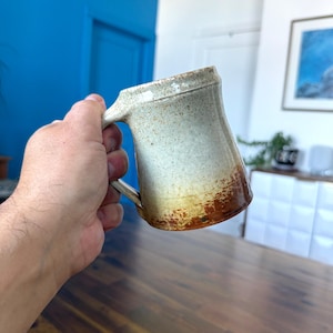 May include: A handmade ceramic mug with a unique, rustic design. The mug features a light beige colour with brown and yellow accents near the base. It has a large handle and a slightly flared rim, perfect for enjoying tea or coffee.
