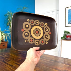 May include: A rectangular brown ceramic tray with rounded corners. The tray features a central design of concentric circles in gold, orange, and brown. The tray is held up by a person's hand, showcasing the decorative pattern.