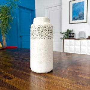 May include: A cream-colored ceramic vase with a cylindrical shape. The upper portion features a pattern of raised, circular designs. The vase is displayed on a dark wooden surface, with a blue door and artwork in the background.