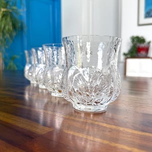 May include: Five clear glass tumblers with a textured thistle design. The glasses are arranged on a wooden surface. The background features a blue door, a white door, and a framed picture.