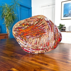 May include: Abstract ceramic art piece with an organic shape. The artwork features red, orange, gold, and white colours, creating a textured surface. The piece is displayed on a wooden table.