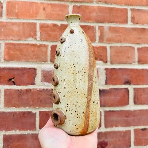 May include: A speckled white ceramic vase with brown accents and a series of holes along the side. The vase has a rounded bottom and a narrow neck.