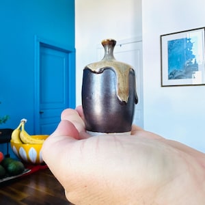May include: A small, handcrafted ceramic vase with a dark brown base and a lighter, dripped glaze around the top. The vase is held in a hand, showcasing its unique shape and texture. The vase is approximately 10 cm tall.