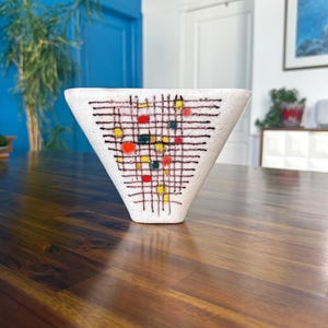 May include: A white ceramic vase with a wide, triangular top and a narrow base. The exterior features a dark brown grid pattern overlaid with small, scattered circular accents in vibrant shades of red, yellow, and dark green.