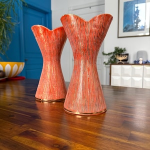 May include: Two orange vases with a textured, vertical stripe design and gold trim. The vases have a unique, flared top and an hourglass shape. They are displayed on a dark wooden table, showcasing a mid-century modern aesthetic.