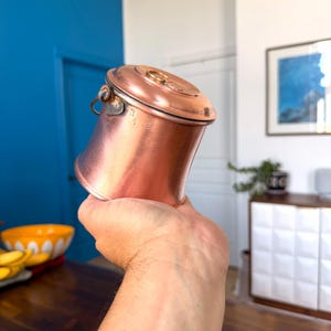 May include: A copper-colored, small pot with a lid and a handle. The pot has a rounded body and a lid with a circular handle. The pot is held in a hand. The background includes a blue wall and a white cabinet.