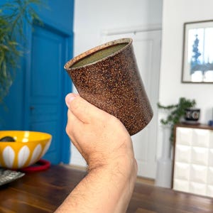 May include: A handmade, speckled brown ceramic jug with a slightly flared rim. The interior has a green glaze. The jug is held in a person's hand. The background includes a blue door and a yellow and white bowl.