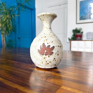 May include: A ceramic vase with a rounded body and a flared neck, featuring a cream-coloured glaze with brown speckles throughout. A single, dark brown, leaf-shaped design is centred on the front of the vase, which has a smooth, glossy finish.