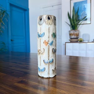 May include: A cylindrical ceramic vase with a speckled off-white glaze. The surface features vertical incised lines and small, hand-painted floral motifs in shades of blue, green, and orange. The top rim has several small circular openings.