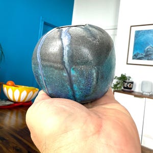 May include: A small, round, blue and grey ceramic vase with a speckled glaze. The vase has a wide opening and is perfect for displaying a single flower or small bouquet.