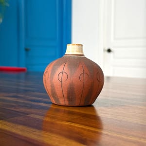 May include: A terracotta-coloured vase with a cream rim and a brown, vertical-line pattern. The vase has a rounded shape and sits on a wooden surface. The background features a blue door and a white door.