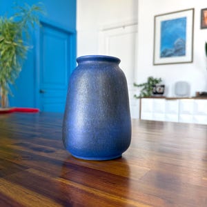 May include: A blue ceramic vase with a textured surface. The vase has a rounded body and a slightly flared rim. The vase is sitting on a wooden table.