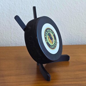 May include: A black hockey puck with the Chicago Blackhawks logo, displayed on a black wooden stand. The puck is round and has the team's logo in the center. The stand has three legs and supports the puck at an angle.