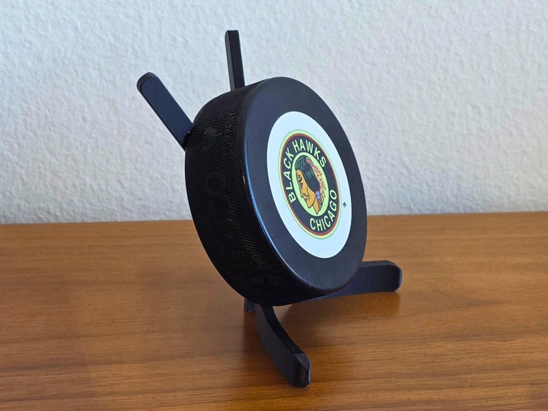 May include: A black hockey puck with the Chicago Blackhawks logo, displayed on a black wooden stand. The puck is round and has the team's logo in the center. The stand has three legs and supports the puck at an angle.