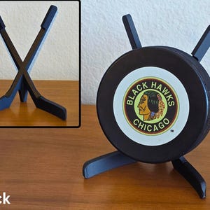 May include: Black hockey puck display stand featuring the Chicago Blackhawks logo. The stand is made of black wood and holds the puck upright. The stand has a unique design with two crossed hockey sticks.