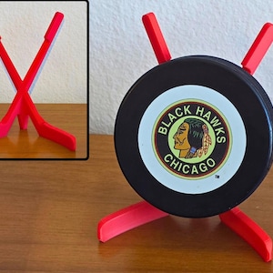 May include: A red hockey puck holder with a black puck featuring the Chicago Blackhawks logo. The holder is made of red plastic and has a unique design to display the puck. The word "Red" is at the bottom.