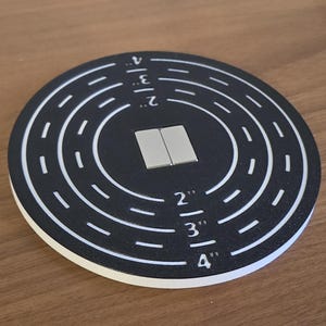 Puede incluir: Un objetivo en blanco y negro con círculos concéntricos y medidas en pulgadas. Dos pequeños imanes rectangulares plateados están en el centro. El objetivo tiene medidas de 2", 3" y 4".