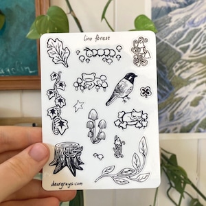 Puede incluir: Una hoja de pegatinas blanca con ilustraciones en negro de elementos temáticos del bosque. Los diseños incluyen una oruga, setas, un pájaro, un gato, hojas y el texto "lino forest" y "deargrays.com".
