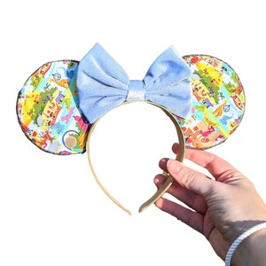 Puede incluir: Diadema con lazo azul claro y tela estampada de colores en las orejas. El estampado presenta un mapa de un parque de atracciones con varias atracciones y personajes.