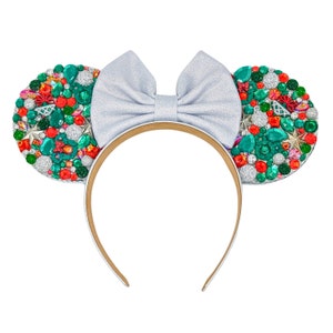 May include: A festive headband with mouse ears, adorned with a silver bow and embellished with colorful gems. The ears feature a mix of red, green, and silver jewels, creating a sparkling effect. The headband is white with a tan interior.