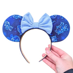 Puede incluir: Orejas de ratón azules y blancas con un lazo azul claro. Las orejas tienen un fondo azul oscuro con siluetas blancas de personajes de Disney.