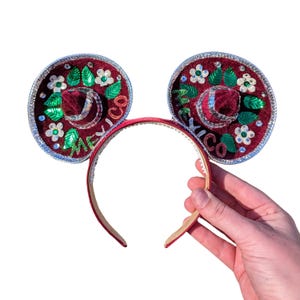May include: Red velvet Mickey Mouse ears with a silver trim and a sombrero design. The sombrero is decorated with green leaves, white flowers, and silver sequins. The word "Mexico" is written on the sombrero.
