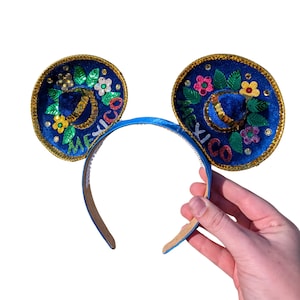 May include: Blue and gold Mickey Mouse ears with a sombrero design. The ears are decorated with colorful flowers, green leaves, and gold glitter. The headband is blue. The word "Mexico" is written on the ears.