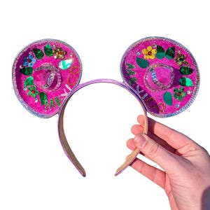 Puede incluir: Orejas de Minnie Mouse rosas con un diseño de sombrero rosa con adornos florales verdes y amarillos y la palabra "México" escrita en la parte delantera.