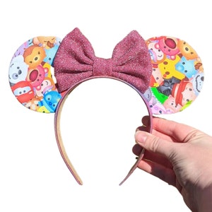 Puede incluir: Una diadema con lazo rosa brillante y dos orejas circulares con un colorido estampado de personajes de dibujos animados. La diadema está sujeta por una mano sobre un fondo blanco. El lazo y las orejas son el punto focal.