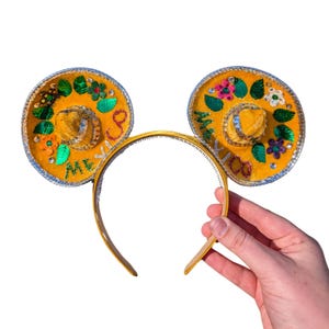 Puede incluir: Orejas de Mickey Mouse de fieltro amarillo con un diseño de sombrero. Las orejas están decoradas con hojas verdes, flores y purpurina. La diadema es dorada. El texto "México" está escrito en las orejas.