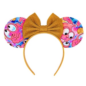 May include: Gold headband with a large gold bow and two pink and blue ears featuring a cartoon character with a brain.