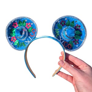 Puede incluir: Diadema de orejas de Mickey Mouse azul con diseño de sombrero. Las orejas están decoradas con flores coloridas, hojas y purpurina. La diadema dice "México" en cada oreja.