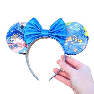 Peut inclure: Un serre-tête bleu avec des oreilles de souris et un nœud en satin. Les oreilles sont décorées d'une scène océanique bleue et de personnages d'un film d'animation populaire. Le serre-tête est tenu par une main.