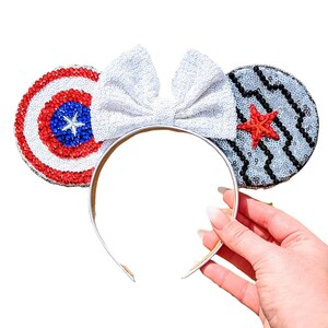 May include: A pair of white sparkly Minnie Mouse ears with a red, white, and blue Captain America shield on one ear and a black and white Captain America shield on the other ear.