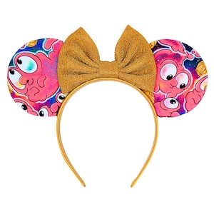 May include: Gold headband with a large gold bow and pink ears featuring a cartoon character with multiple eyes.