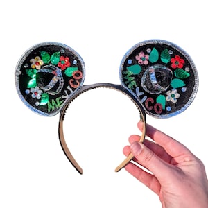 Puede incluir: Diadema negra con dos orejas de fieltro negro decoradas con purpurina plateada y bordados coloridos. Las orejas tienen forma de sombreros con la palabra "México" escrita en ellas.