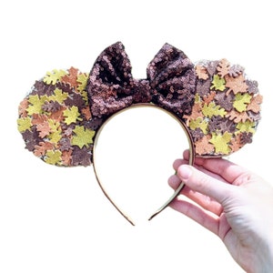 Puede incluir: Un par de orejas de ratón marrones y doradas con un lazo de lentejuelas marrones. Las orejas están cubiertas de pequeñas hojas brillantes.