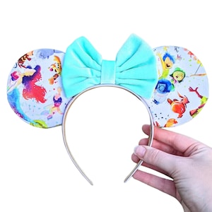 Könnte beinhalten: Ein Paar hellblaue Mäuseohren mit einer hellblauen Samtschleife. Die Ohren sind mit einem bunten Druck von Disney-Charakteren verziert, darunter Arielle, Tinkerbell und die Figuren aus Alles steht Kopf.