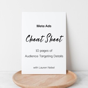 Puede incluir: Una "Meta Ads Cheat Sheet" blanca con texto negro, que incluye "10 páginas de detalles de segmentación de audiencia" y el nombre del autor, descansa sobre una bandeja de madera marrón claro. El fondo es una superficie blanca lisa.