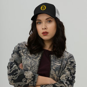 May include: A woman wearing a black and white trucker hat with a yellow Bitcoin symbol embroidered on the front. She is wearing a black and gray patterned jacket and has her arms crossed.