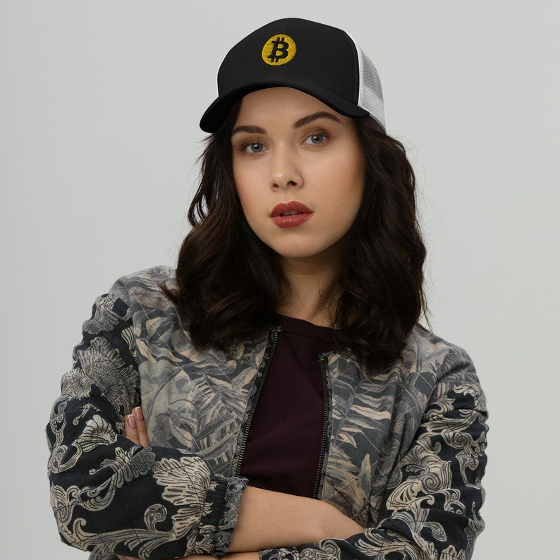 May include: A woman wearing a black and white trucker hat with a yellow Bitcoin symbol embroidered on the front. She is wearing a black and gray patterned jacket and has her arms crossed.