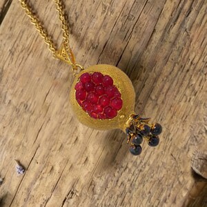 May include: A gold-colored necklace with a pendant shaped like a pomegranate. The pendant features a textured gold exterior and a cluster of red gems representing the seeds. Small blue gems are at the base.