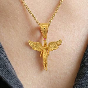 May include: A gold-colored angel pendant necklace. The pendant features an angel with outstretched wings and a small red gemstone. The necklace is worn on a person wearing a dark gray shirt.